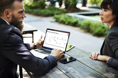A man and a woman engage in a serious discussion at an outdoor table. The man, with a long beard, shows the woman something on his laptop screen, which displays the Purchase Only | No Online Access Innovation Prioritization Tool & Dashboard.